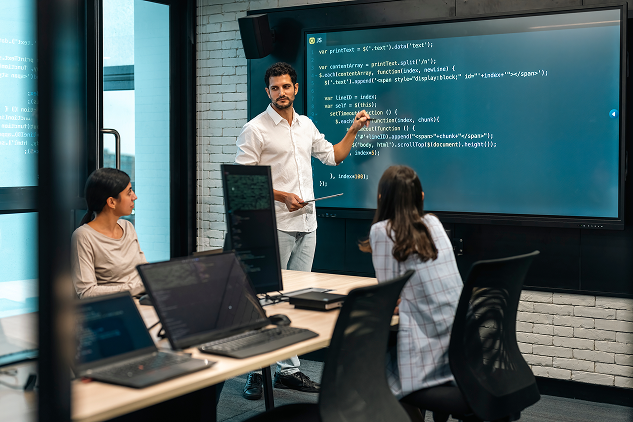 Man pointing to code on a large display in a modern office setting. Two women are sitting and looking at the display.