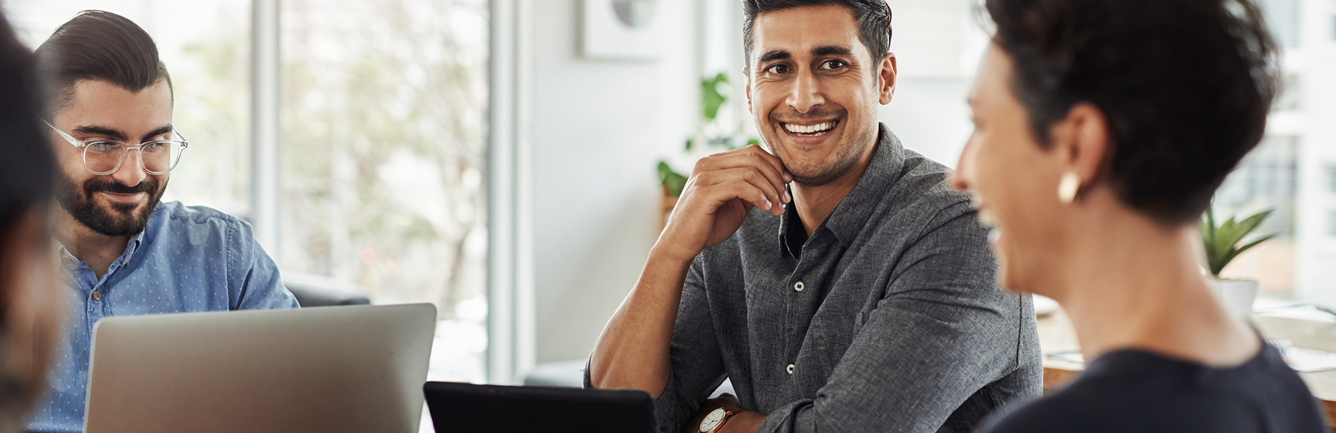 A diverse group of professionals collaborate at a bright, modern office. One man smiles directly at the camera. Laptops and a tablet are visible