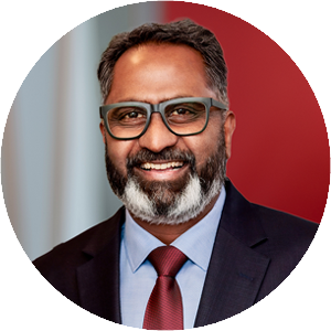 Man (Natarajan Venkatesh) smiling in front of a modern office background with red and white accents wearing glasses, a beard, and a suit and tie.