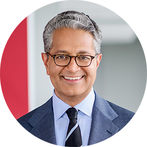 A smiling man (Salim Ramji) with gray hair and glasses, clad in a blue suit and a black and white tie, is set against a red and gray backdrop.