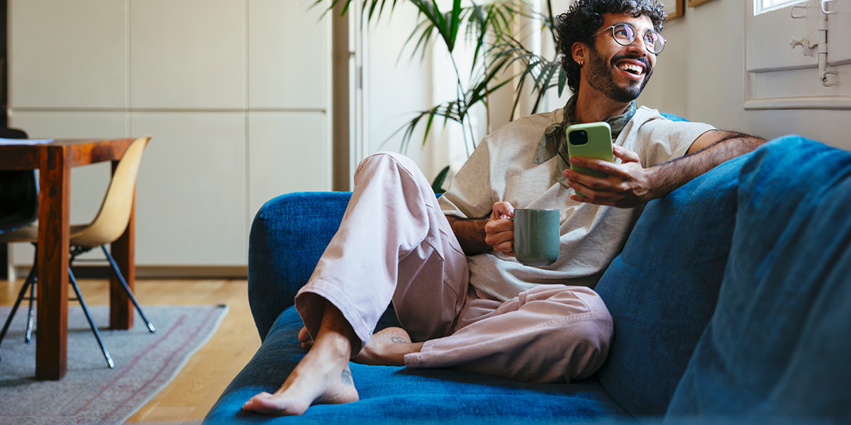Man smiling and sitting on a sofa holding a mobile phone in his left hand and coffe cup in his right hand.